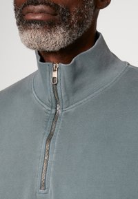 Lichtblauwe kwartzip pullover, gemaakt van een textuurstof. Heeft een zilveren rits met een ronde trekker. Close-up toont de halslijn en het patroon.