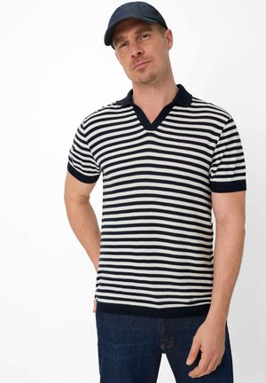 Man met een marineblauwe pet, een zwart-wit gestreept poloshirt met korte mouwen en donkerblauwe spijkerbroek, staand met één hand achter zijn rug.