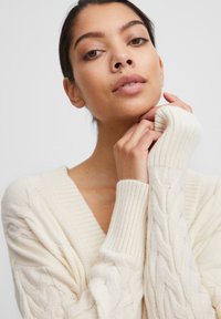 Cremiger, gestrickter Pullover mit tiefem V-Ausschnitt, gerippten Bündchen und strukturierten Zopfmustern. Weicher Stoff mit einer gemütlichen, lässigen Passform.