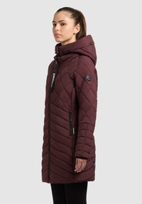 Femeie purtând o geacă cusută maro închis cu glugă și buzunare cu fermoar, stând lateral pe un fundal simplu, gri deschis.