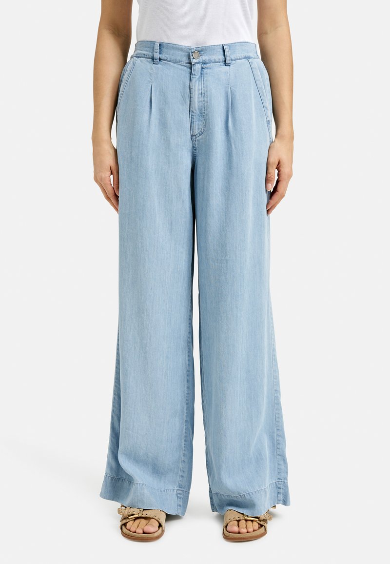 Light blue, wide-leg denim trousers with pleats at the waist, a button closure, and contrasting stitching; paired with tan sandals.