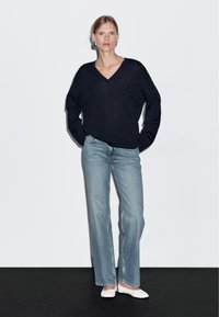 Marinblå stickad tröja med V-ringning och avslappnad passform. Parat med ljusblå raka jeans och vita ballerinaskor. Modellen står framför en neutral bakgrund.