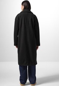 Lungo cappotto di lana nera con ampio colletto e spalle basse, caratterizzato da una linea dritta e fessure laterali all'orlo, indossato sopra jeans blu.