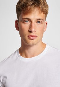 Hvid bomulds T-shirt med rund hals, med korte ærmer og en glat tekstur, båret af en mandlig model mod en neutral baggrund.