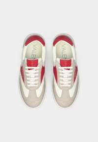 Ζευγάρι λευκά και κόκκινα παπούτσια Valentino με μπεζ σουέντ λεπτομέρειες και λευκά κορδόνια, απεικονιζόμενα από πάνω σε λευκό φόντο.