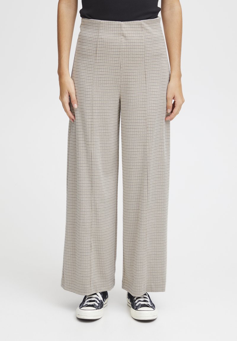 Brede broek in een lichtbeige houndstoothpatroon, met een hoge taille, plooien en een zachte stof. Gecombineerd met zwarte sneakers.