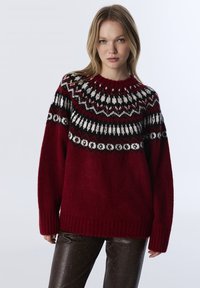 Pull en tricot rouge avec un empiècement orné en noir et blanc. Dispose d'un col rond et de poignets et d'un ourlet côtelés. Texture douce.