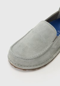 Light gray suede slip-on shoe with rounded toe, stitched detailing, and rubber sole, featuring a textured surface and a blue insole.