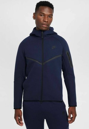 Granatowa kurtka Nike zapinana na zamek z kapturem, z czarnym ukośnym akcentem, kieszeniami po bokach oraz eleganckim, teksturowanym wykończeniem.