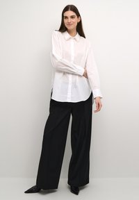 Camicia bianca con colletto texture e maniche lunghe, abbinata a pantaloni neri larghi e scarpe nere con punta.