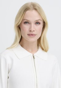 Vêtement en maille blanche avec un col et une fermeture éclair frontale. Le tissu semble lisse et côtelé, mettant en valeur un design minimaliste.