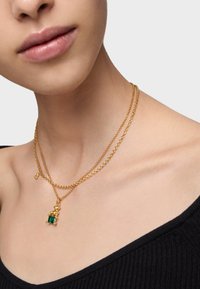 Złota naszyjnik warstwowa z wieloma łańcuszkami; jeden z nich ma zawieszkę z zielonym kamieniem szlachetnym, a inny ma mały urok. Widoczna faktura wykończenia.
