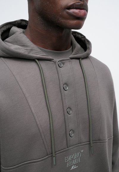 Hanorac gri cu glugă, dotat cu două butoane, sireturi și logo brodat. Țesătură netedă cu cusături texturate și o croială lejeră.