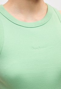 Κοντινό πλάνο ενός ατόμου που φοράει ένα ριμπ ύφασμα αμάνικο τοπ σε ανοιχτό πράσινο χρώμα με ένα μικρό κεντητό λογότυπο στο στήθος.