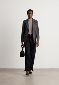 Blazer gris sur un haut beige, associé à un pantalon noir avec des motifs subtils. Le modèle tient un sac à main noir et porte des ballerines noires.