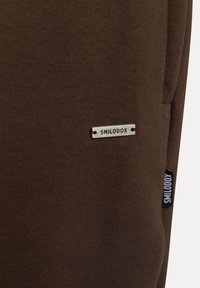 Braune Rippstoff-Jogginghosen mit einem silbernen "SMILODOX"-Label und einem schwarzen "SMILODOX"-Tag, die ein enganliegendes Design und eine glatte Textur hervorheben.