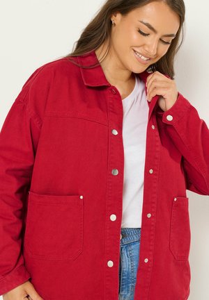 Vrouw die een rood jack met knopen draagt over een wit shirt en blauwe jeans, glimlachend en met één hand de kraag van het jack rechtzettend.