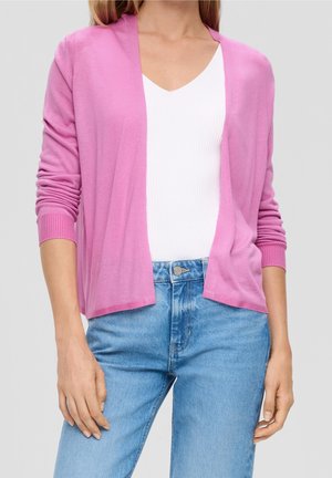 Femme portant un cardigan rose clair sur un haut blanc à col en V et un jean taille haute bleu, debout devant un fond gris uni.