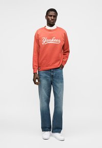 Homme portant un sweat-shirt Yankees corail, un jean bleu clair et des baskets blanches, debout devant un fond blanc uni.