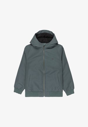 Giacca impermeabile verde con cappuccio, cerniera frontale e tasche laterali, indossata sopra una t-shirt grigia e pantaloni larghi verde oliva.