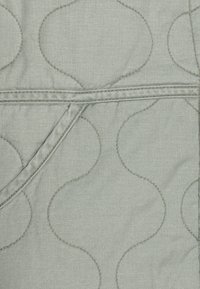Jasnozielona pikowana tkanina z falistym wzorem, z widocznymi liniami szycia i detalem w postaci zagięcia w pobliżu krawędzi. Miękka tekstura.