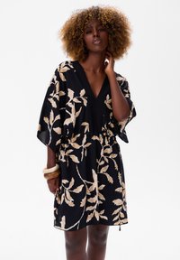 Zwarte jurk met beige bladenpatronen, V-hals, brede mouwen en een elastische taille. Gemaakt van lichtgewicht stof met een zachte textuur.