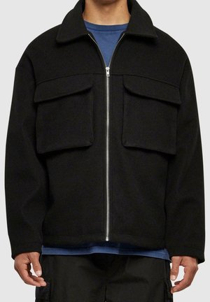 Mann trägt schwarze Reißverschlussjacke mit großen Klappentaschen, darüber ein blaues Hemd, Hände locker an den Seiten.