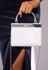 Fehér táska strukturált doboz formával, ezüst szegecsekkel díszítve és elegáns ezüst fogantyúval. Levehető ezüst láncos pánttal.