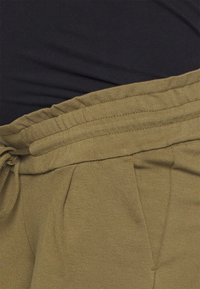 Olivové běžecké kalhoty s tkaničkou v pase, které mají příjemnou texturu, sklady a elastický pas. Černá látka viditelná nahoře.