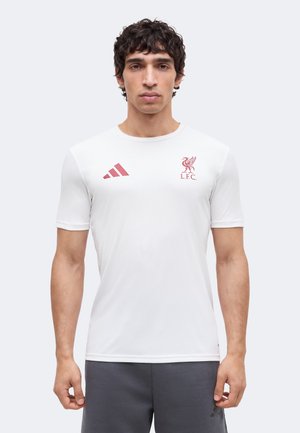 Homme portant un maillot de sport blanc du Liverpool FC avec des logos rouges et un short gris foncé, debout devant un fond uni.