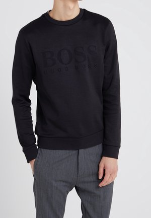 Mężczyzna w czarnej bluzie Hugo Boss z tłoczonym logo i szarych spodniach w prążki, stojący z jedną ręką w kieszeni.