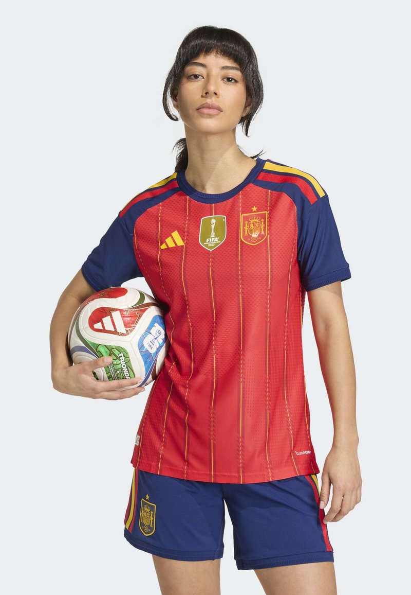 Red and blue soccer kit featuring horizontal stripes, gold accents, and logos. Player holds a multicolored soccer ball in one hand.