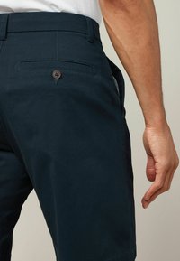Pantalones cortos de un azul marino oscuro, confeccionados en una tela suave, que presentan un bolsillo trasero con un detalle de botón marrón y una abertura lateral, exhibidos en una mano.