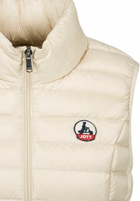 Gilet rembourré beige clair avec un col montant, fermeture éclair sur le devant et un patch en forme de rond avec un cycliste, motif matelassé texturé sur l'ensemble.