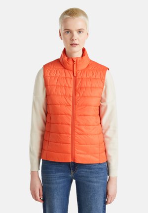 Oranje gewatteerde vest met een hoge kraag en een rits aan de voorkant. Draagt over een lichtgekleurde lange mouw shirt, gecombineerd met blauwe jeans.