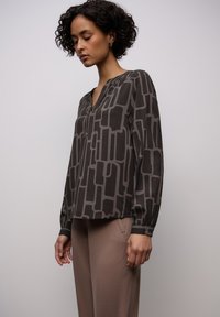 Blusa a maniche lunghe di colore grigio scuro con motivo geometrico in grigio più chiaro. Presenta una scollatura a V e polsini arricciati. Tessuto liscio e leggero.