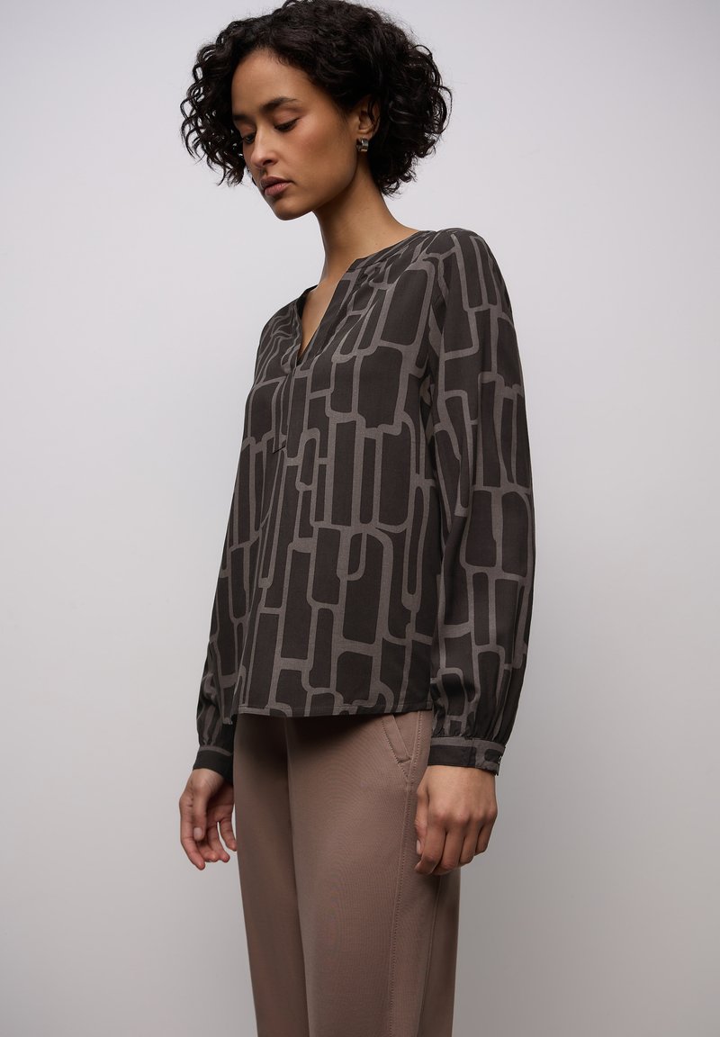 Blusa a maniche lunghe di colore grigio scuro con motivo geometrico in grigio più chiaro. Presenta una scollatura a V e polsini arricciati. Tessuto liscio e leggero.