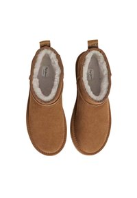 Botas de tobillo de ante marrón con un suave forro de piel blanca, punta redondeada y tiradores a los lados para ponerlas y quitarlas fácilmente. Diseño de suela plana.