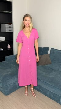 Γυναίκα που στέκεται μέσα σε εσωτερικό χώρο, φοράει ροζ φόρεμα με κουμπιά και λευκά πέδιλα με τακούνι, δίπλα σε μπλε καναπέ και μαύρη βιβλιοθήκη.