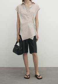 Camicia avvolgente beige con righe e tasca, pantaloni corti neri, borsa a tracolla nera e infradito neri. Tessuto morbido e design minimalista.
