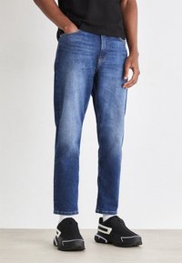 Jeans de mezclilla en azul oscuro con corte ajustado, ligero desgastado, diseño de cinco bolsillos y detalles cosidos, combinados con zapatillas deportivas negras.