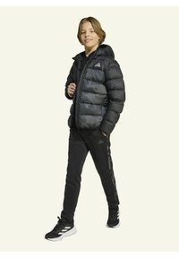 Veste puffer noire avec un design camouflage, fermeture éclair et capuche. Associée à un pantalon de sport noir et des baskets noires avec des accents blancs.