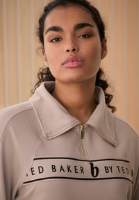 Hellbeige Zip-Up-Oberteil mit gerippter Textur. Verfügt über einen hohen Kragen und ein schwarzes Logo-Band über der Brust. Mit Reißverschluss vorne.