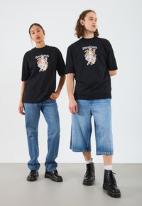 To personer har på seg grafiske T-skjorter i svart med pastellfarger, kombinert med lyseblå denimjeans og svarte snøresko.