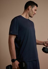 Homme en tenue de sport bleu marine tenant des haltères de 5 kg dans chaque main, sur un fond beige uni.