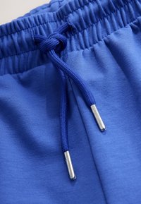 Kék sport rövidnadrág rugalmas derékpánttal és húzózsinórral. A húzózsinóron fém végzárók találhatók. Simaszerű anyagtextúra minták nélkül.