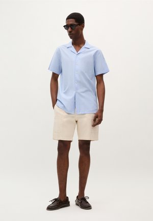 Chemise à manches courtes rayée bleu clair avec un col, short beige et chaussures bateau marron foncé. Portée avec des lunettes de soleil noires.