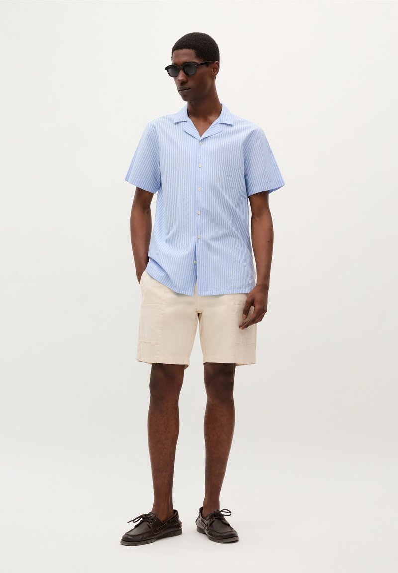 Chemise à manches courtes rayée bleu clair avec un col, short beige et chaussures bateau marron foncé. Portée avec des lunettes de soleil noires.