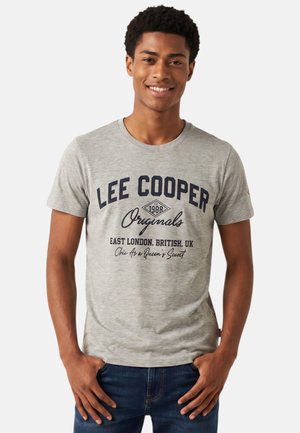 Jeune homme souriant, portant un t-shirt gris Lee Cooper Originals et un jean bleu, debout les mains dans les poches sur fond blanc.