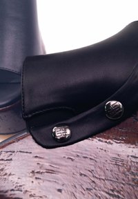 Cavigliera in pelle nera con una vestibilità aderente, caratterizzata da due bottoni metallici e dettagli elasticizzati su una superficie di legno. Texture liscia, design elegante.